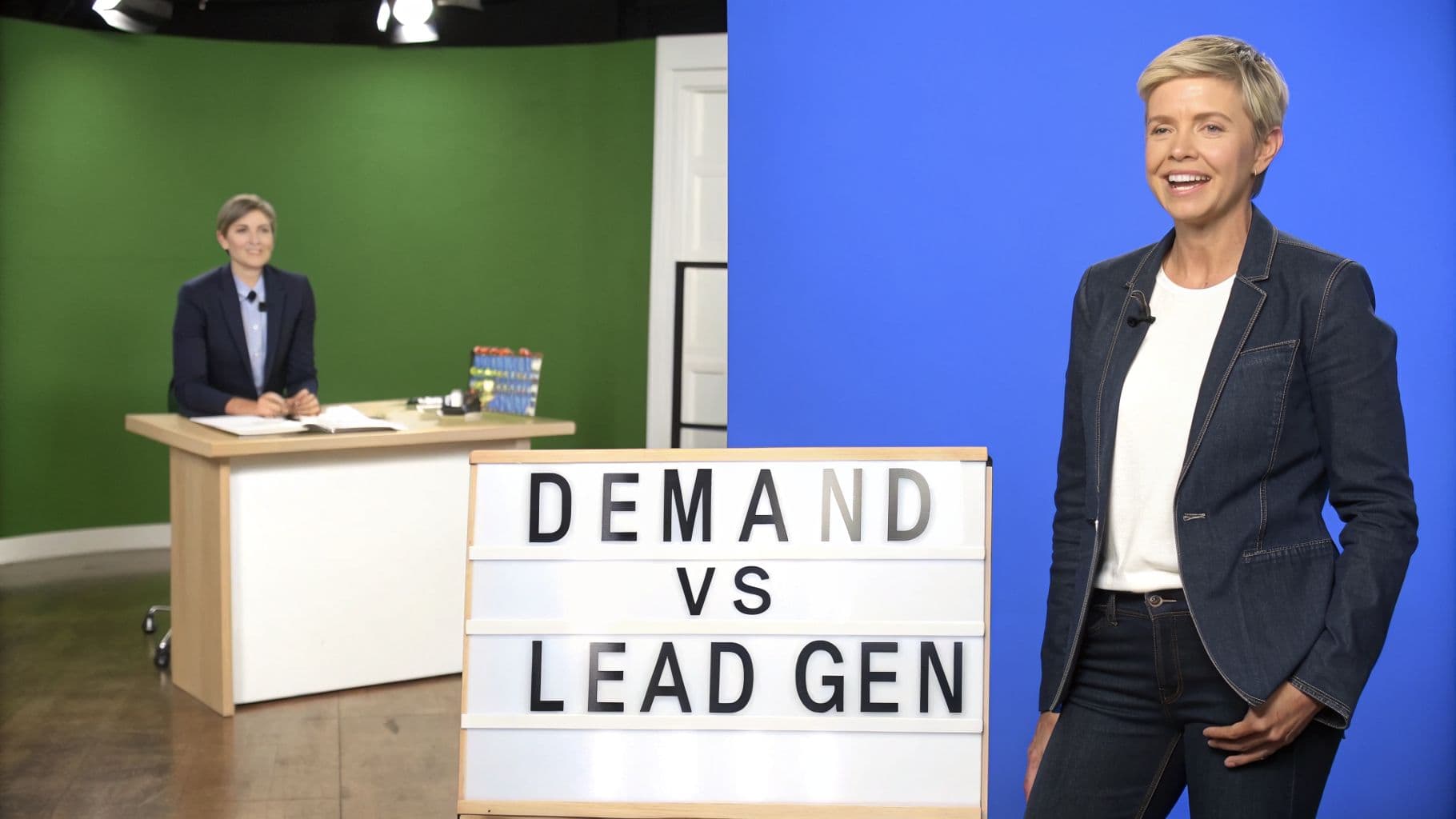 Two women in a studio, one at a desk and one standing, next to a 'Demand vs Lead Gen' sign.