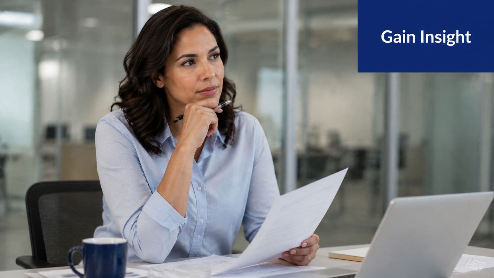 A professional woman reading a document while sitting at her office desk with a laptop.