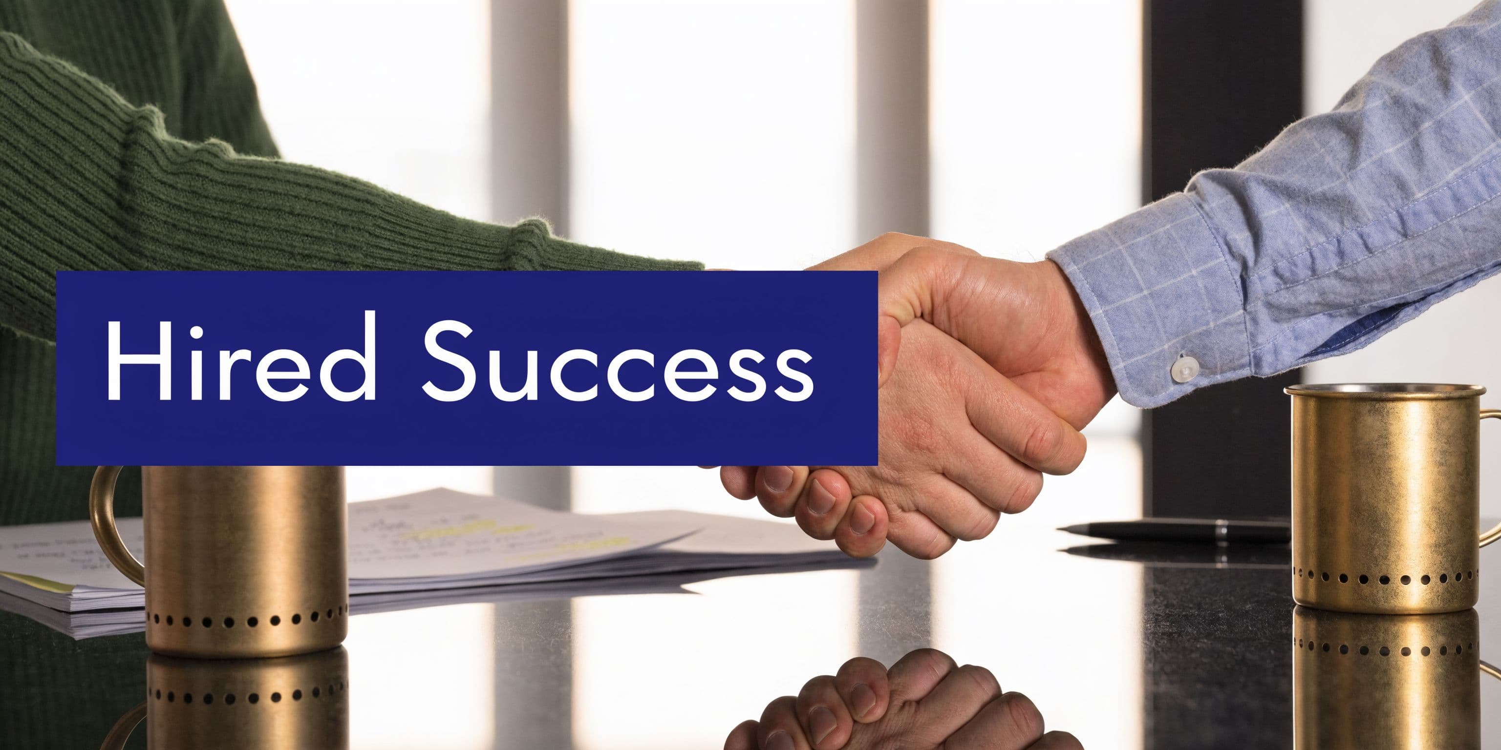 Two professional people shaking hands over a desk, symbolizing a successful recruitment or business hiring partnership.