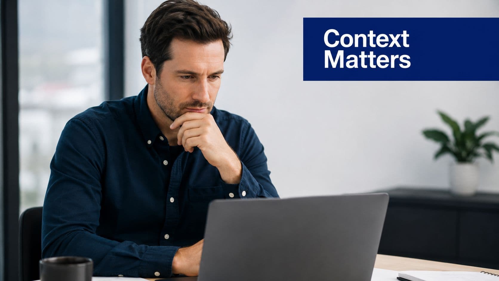 A focused man sitting at a desk and thinking while working on his laptop in an office.