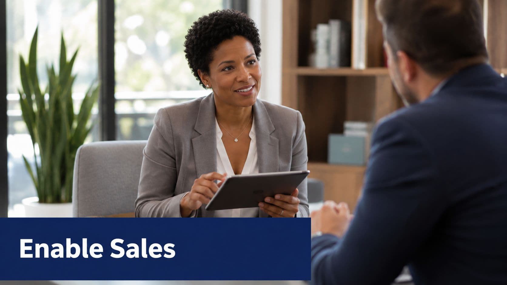 A professional woman in a grey suit discusses strategies on a tablet with her male colleague.