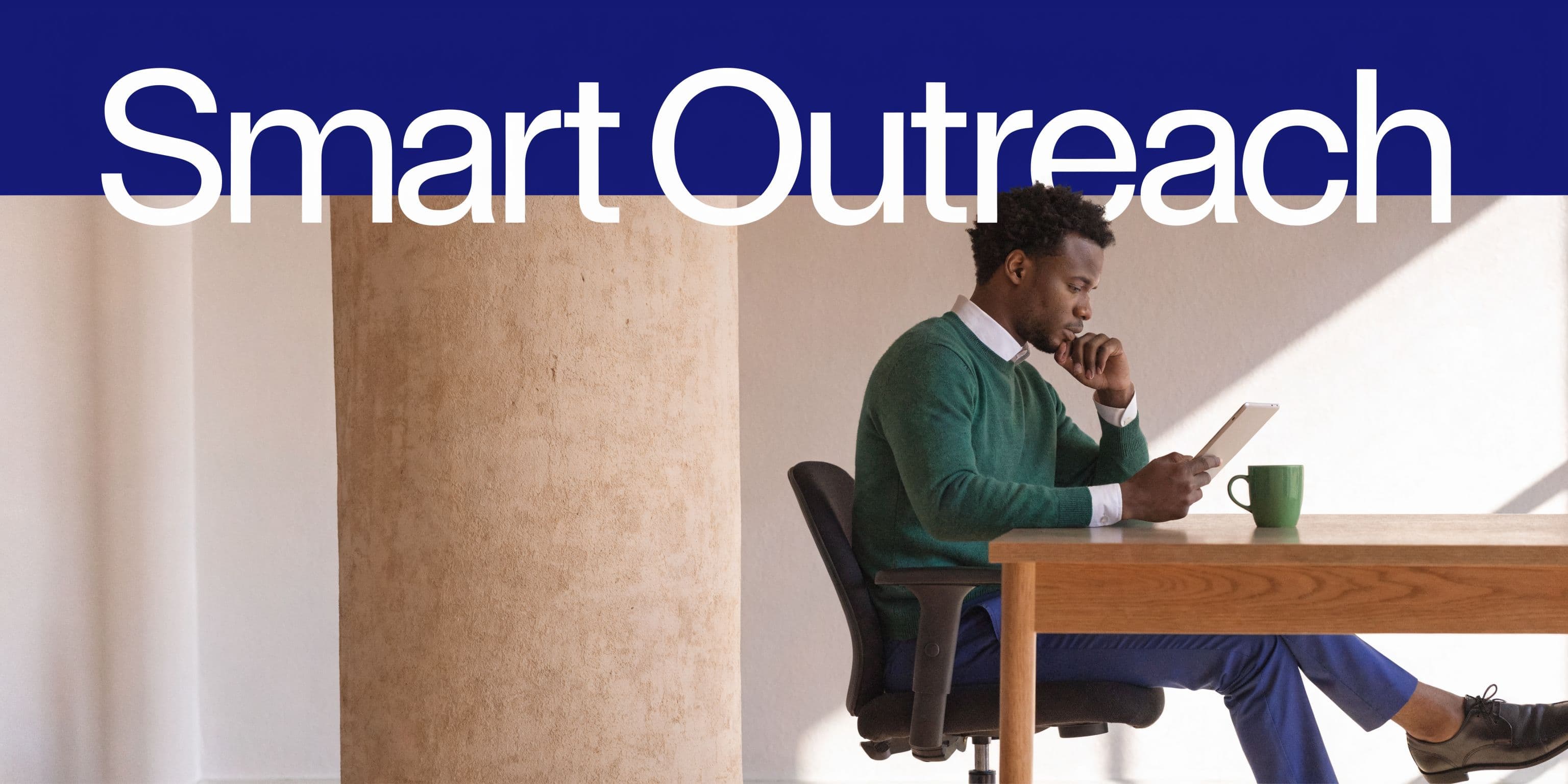 A professional man in a green sweater sits at a desk using a tablet with a cup nearby.