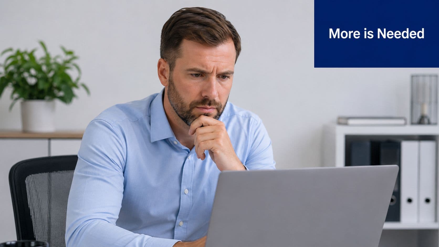 A focused professional man sitting at his desk looking at a laptop computer with a pensive expression.
